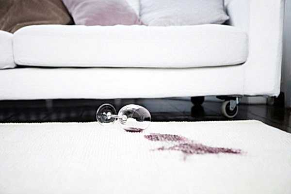 a wine glass is tipped over on a white carpet, with a red wine stain visible. a white sofa is in the background.