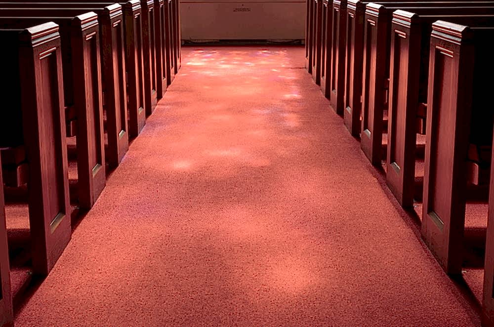 sunlight filters onto a red carpeted aisle between wooden pews inside a church, creating small patches of light on the floor.