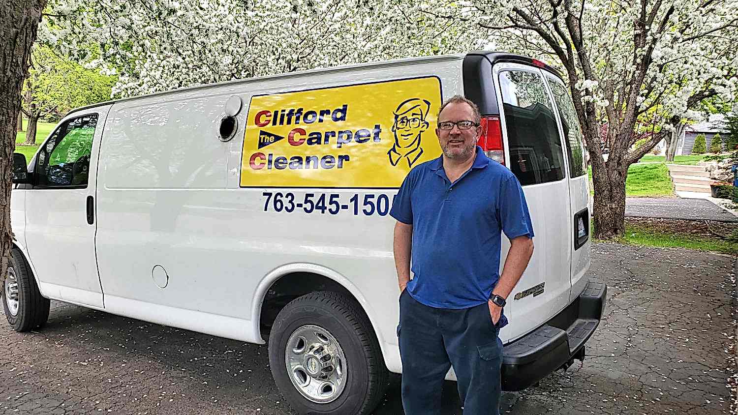 Clifford Cleaning Living Room a man stands in front of a white van with a "clifford the carpet cleaner" sign, parked on a paved driveway under blooming trees.