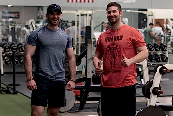 two men pose and smile in a gym; one wears a gray shirt and black shorts, the other a red t shirt with graphics and black shorts, both standing near exercise equipment.